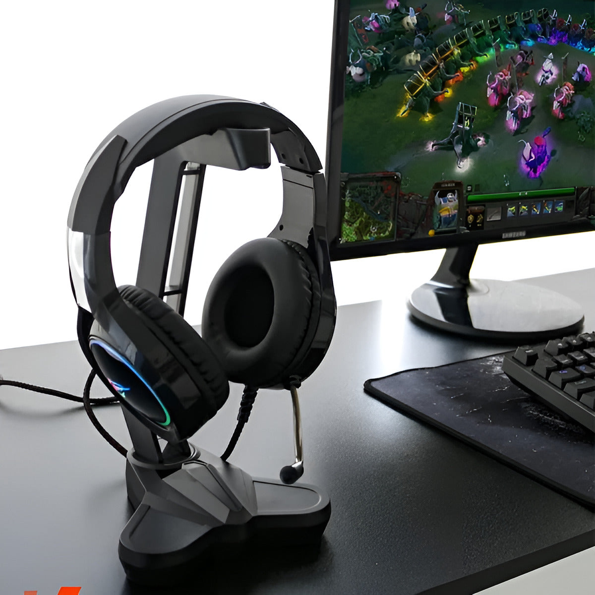 Gaming headset on a stand next to a computer monitor displaying a game, with a keyboard and mouse pad in the foreground.