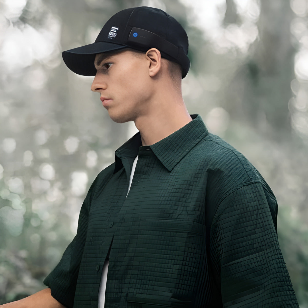 Man wearing a dark green shirt and black cap with a logo, standing outdoors with a blurred natural background.
