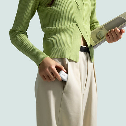 Person wearing a green sweater and beige pants holding a small white device against a light blue background