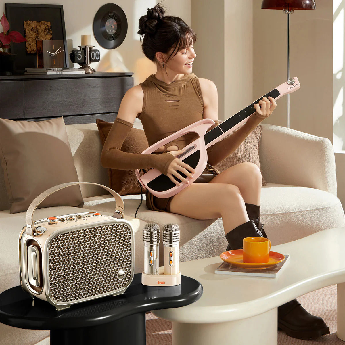 A woman sits on a cream sofa in a living room, holding a pink electric guitar. In the foreground, a small black side table holds a cream portable radio and two matching microphones on a white stand, with a yellow-orange cup on a saucer nearby.