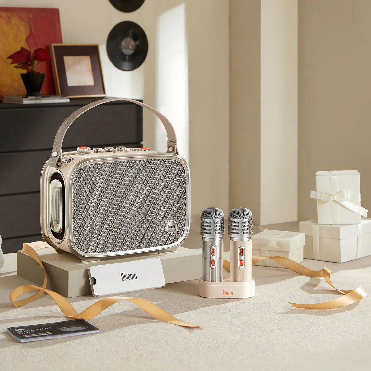 A retro-style portable radio with a handle sits on a table in a styled room setting, accompanied by two small microphones on a stand and gift boxes. The radio has a beige and silver color scheme with a visible 'bloom' brand label.