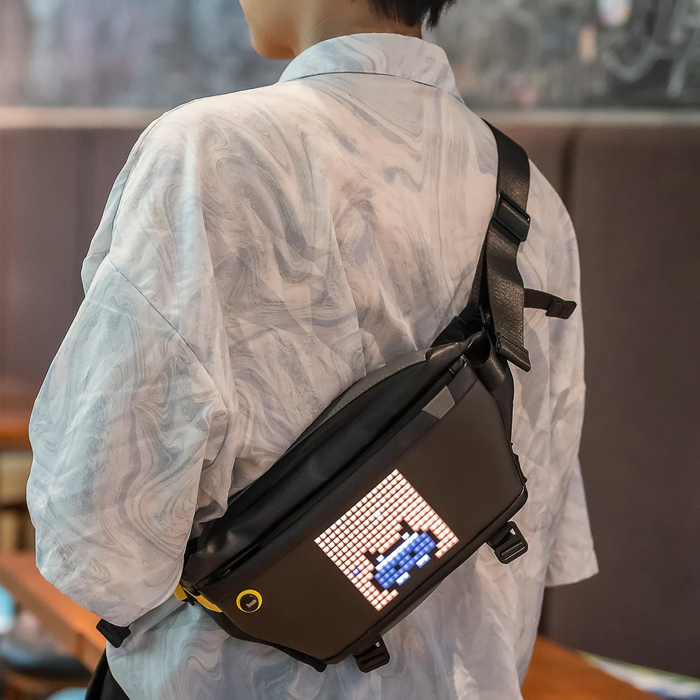 Divoom Pixoo Slingbag‑V Smart LED Sling Bag (4.7" Pixel Screen, App Control) Person wearing a black crossbody bag with a digital display, standing indoors.