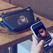 Divoom Pixoo Slingbag‑V Smart LED Sling Bag (4.7" Pixel Screen, App Control) Person using a smartphone with pixel art app on a wooden table