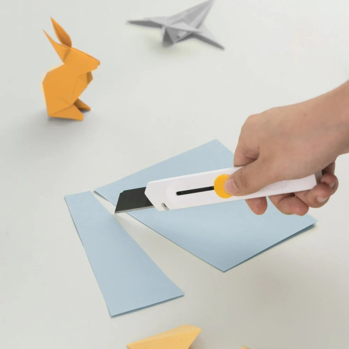 HOTO Precision Utility Knife 18mm Hand using a paper knife to cut blue paper with origami creations in the background.