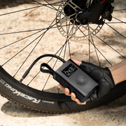 HOTO Electric Air Pump Portable Tire Inflator Hand holding a digital bike pump next to a bicycle wheel on a gravel surface