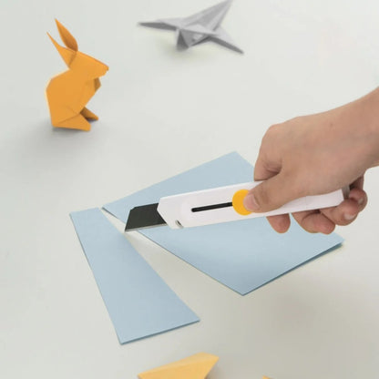 Hand using a paper knife to cut blue paper with origami creations in the background.