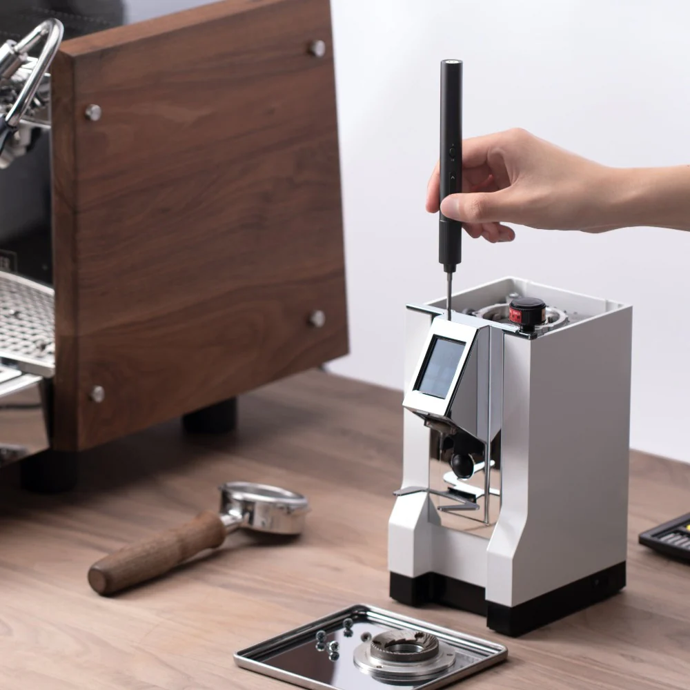 Coffee grinder on a wooden surface with a hand holding a tool, surrounded by coffee-making equipment.