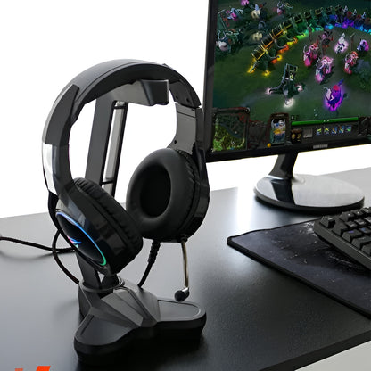 Gaming headset on a stand next to a computer monitor displaying a game, with a keyboard and mouse pad in the foreground.