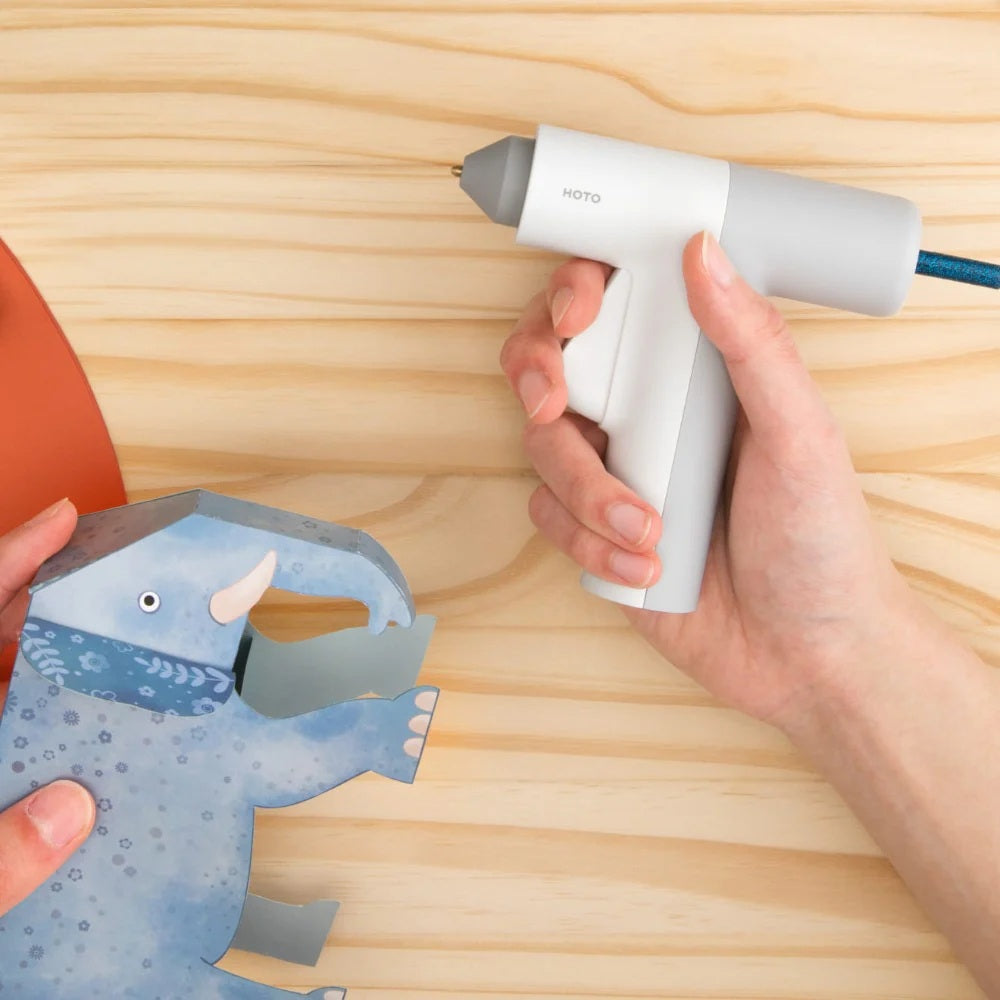 HOTO Lithium Glue Gun, Hand holding a white hot glue gun near a blue paper craft on a wooden surface