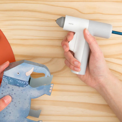 HOTO Lithium Glue Gun, Hand holding a white hot glue gun near a blue paper craft on a wooden surface