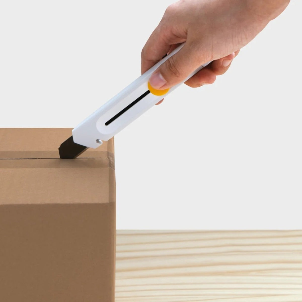 HOTO Precision Utility Knife 18mm Hand using a box cutter to open a cardboard box on a light wooden surface.