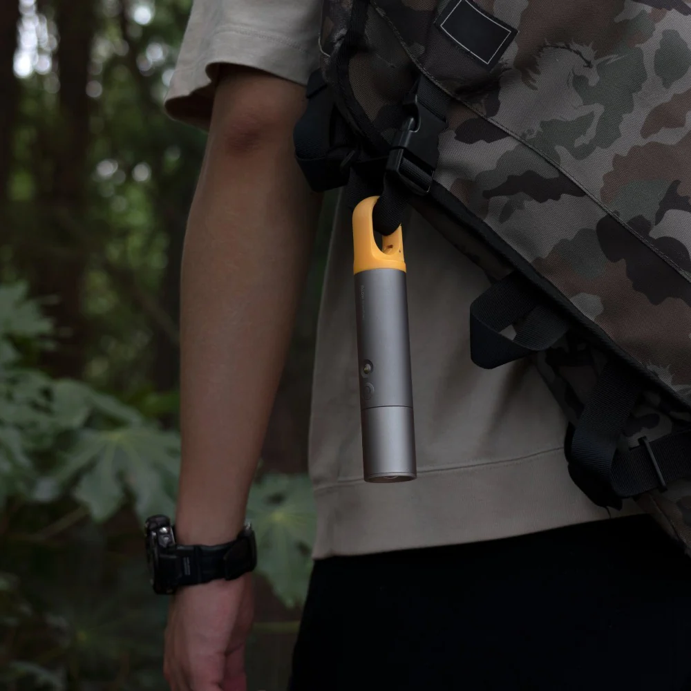 HOTO Flashlight Lite, Person holding a silver and yellow flashlight with a camouflage backpack in a forest setting