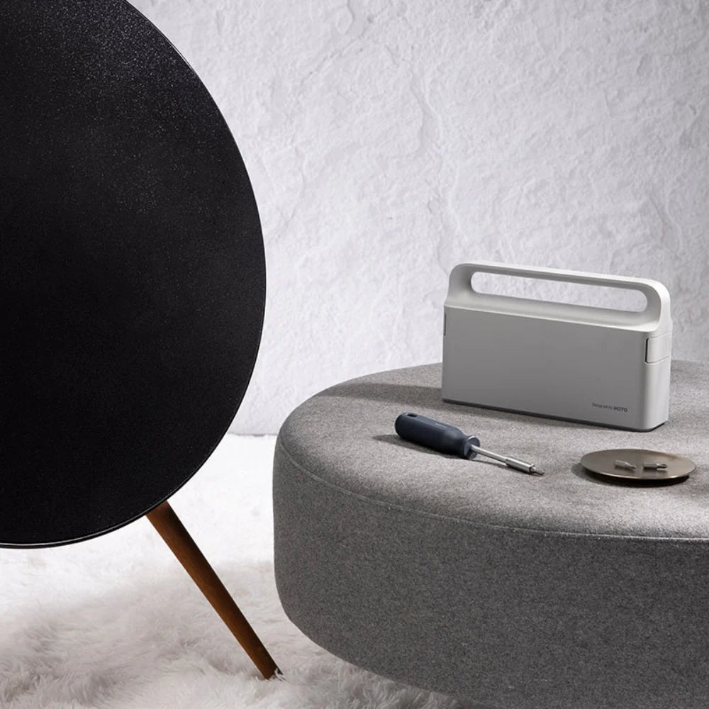White portable speaker on a gray surface with a black spherical object and tools in the background.