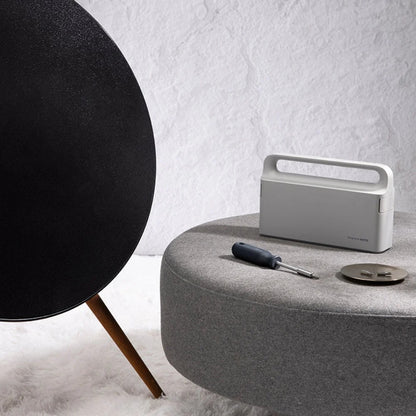 White portable speaker on a gray surface with a black spherical object and tools in the background.
