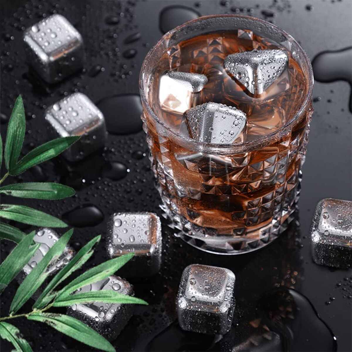 Glass of brown liquid with metal ice cubes on a dark surface with green leaves.