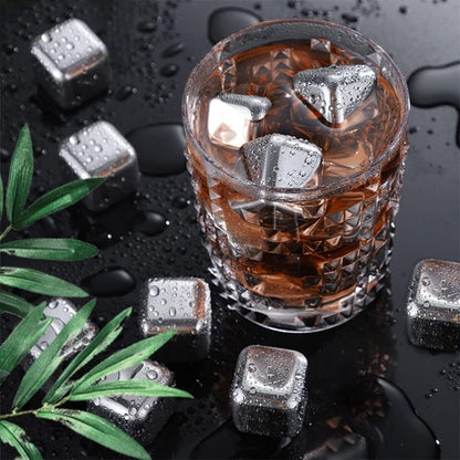 Glass of brown liquid with metal ice cubes on a dark surface with green leaves.